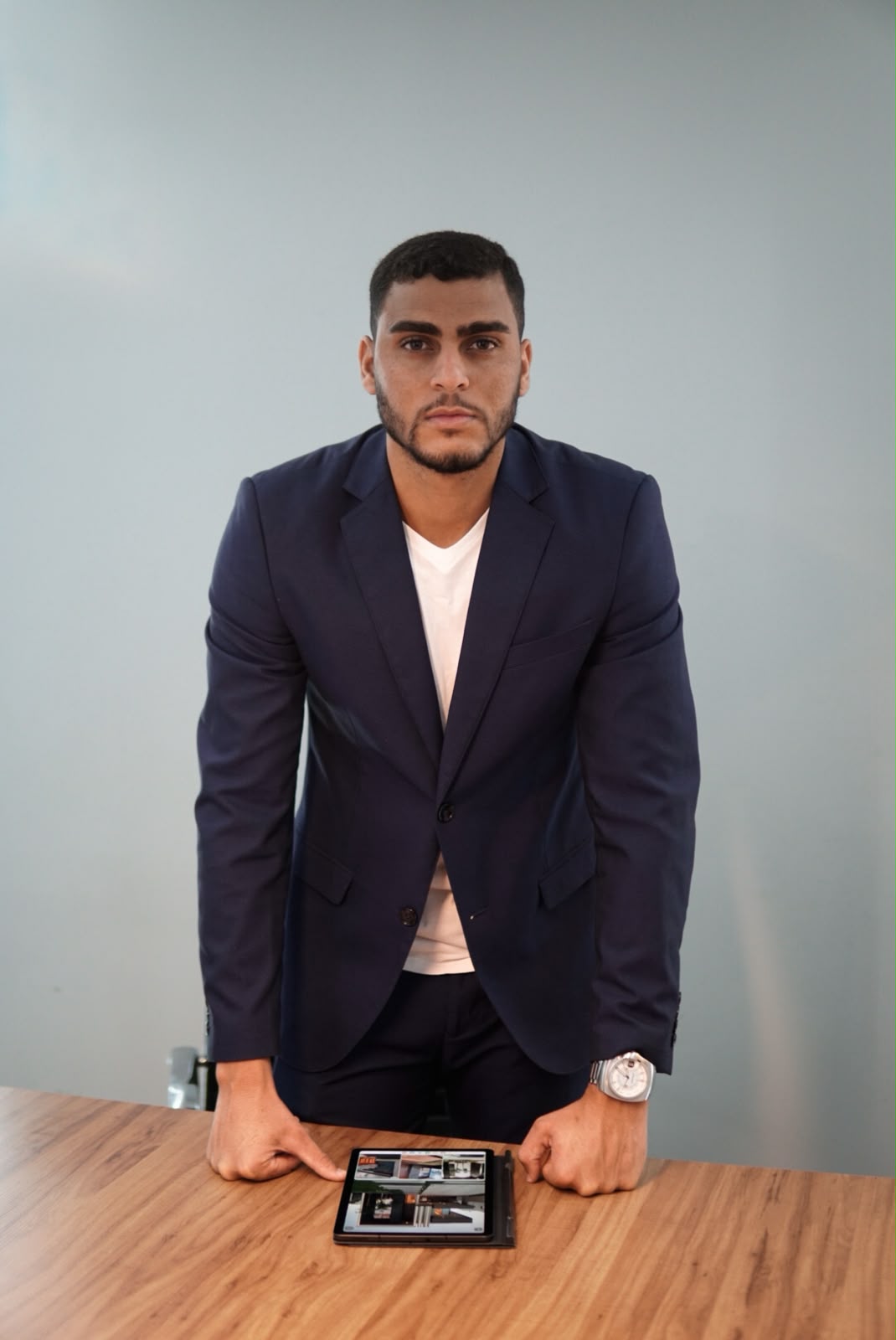 Enzo Reis, à frente da Reis Imóveis, de blazer azul e camiseta branca, em pé com as mãos apoiadas na mesa